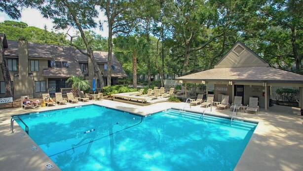 A swimming pool with lounge chairs and a covered pavilion, surrounded by trees and buildings. - Large Golf Course Villa with Lots of Space for Everyone! (Hilton Head)