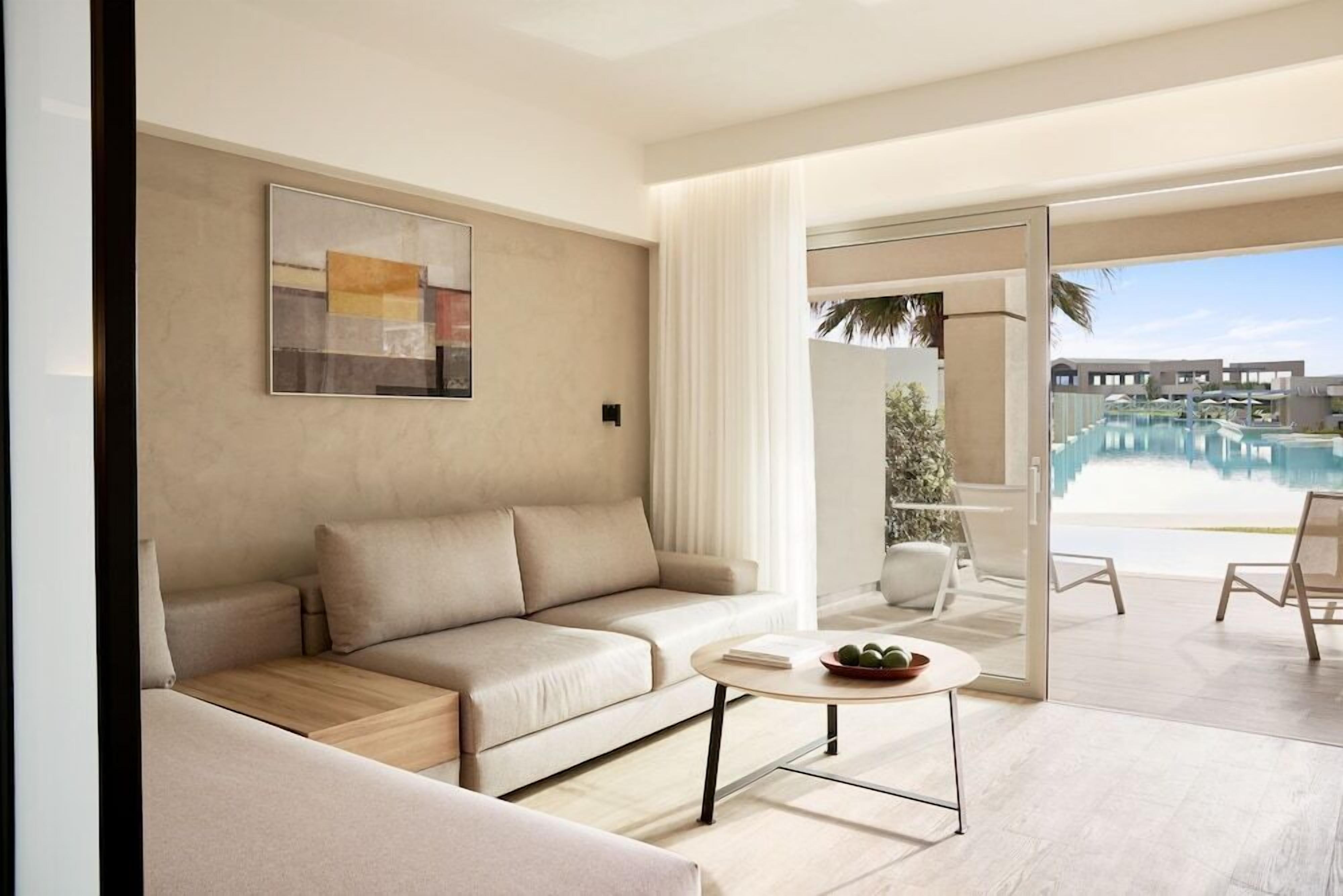 Een moderne woonkamer met een beige bank, een bijzettafeltje met een schaal fruit en een groot raam met uitzicht op een zwembad en resortgebouwen.