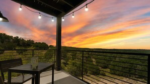 Un balcon avec une table et des chaises, donnant sur un paysage arboré et un coucher de soleil.