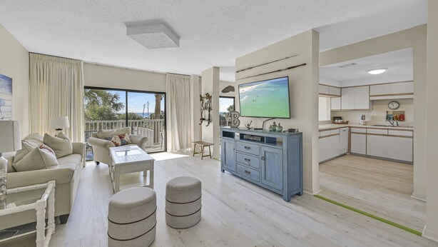 Uma sala de estar moderna com sofá, pufes e TV. Há uma varanda com cortinas e vista para o exterior.