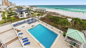 A large swimming pool with lounge chairs, a small building with a green roof, and a beach in the background.