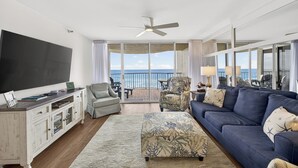 A living room with a large TV, a sofa, a coffee table, and a view of the ocean through large windows.
