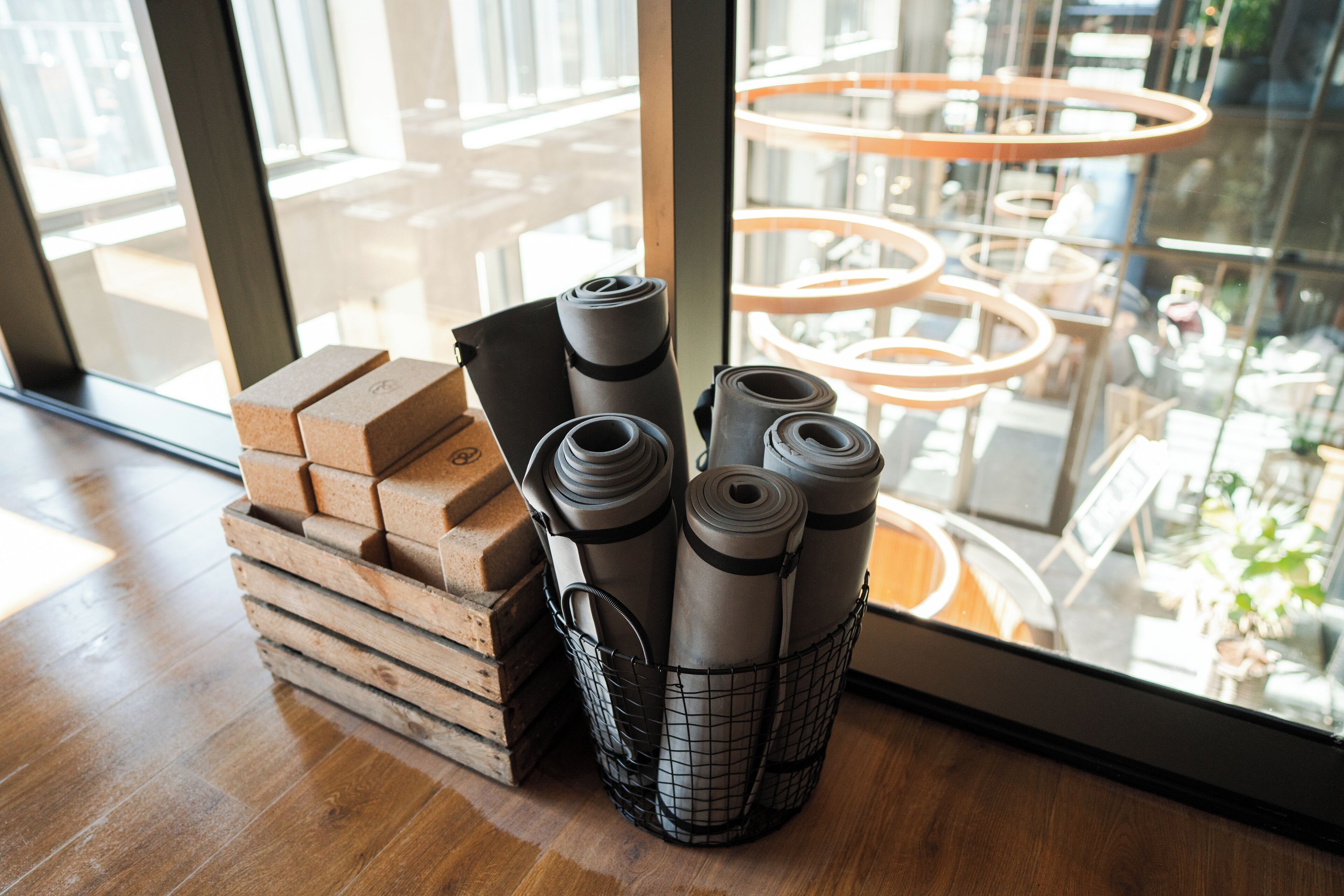 A stack of yoga mats and blocks on a wooden floor next to a window with a view of a modern interior space.
