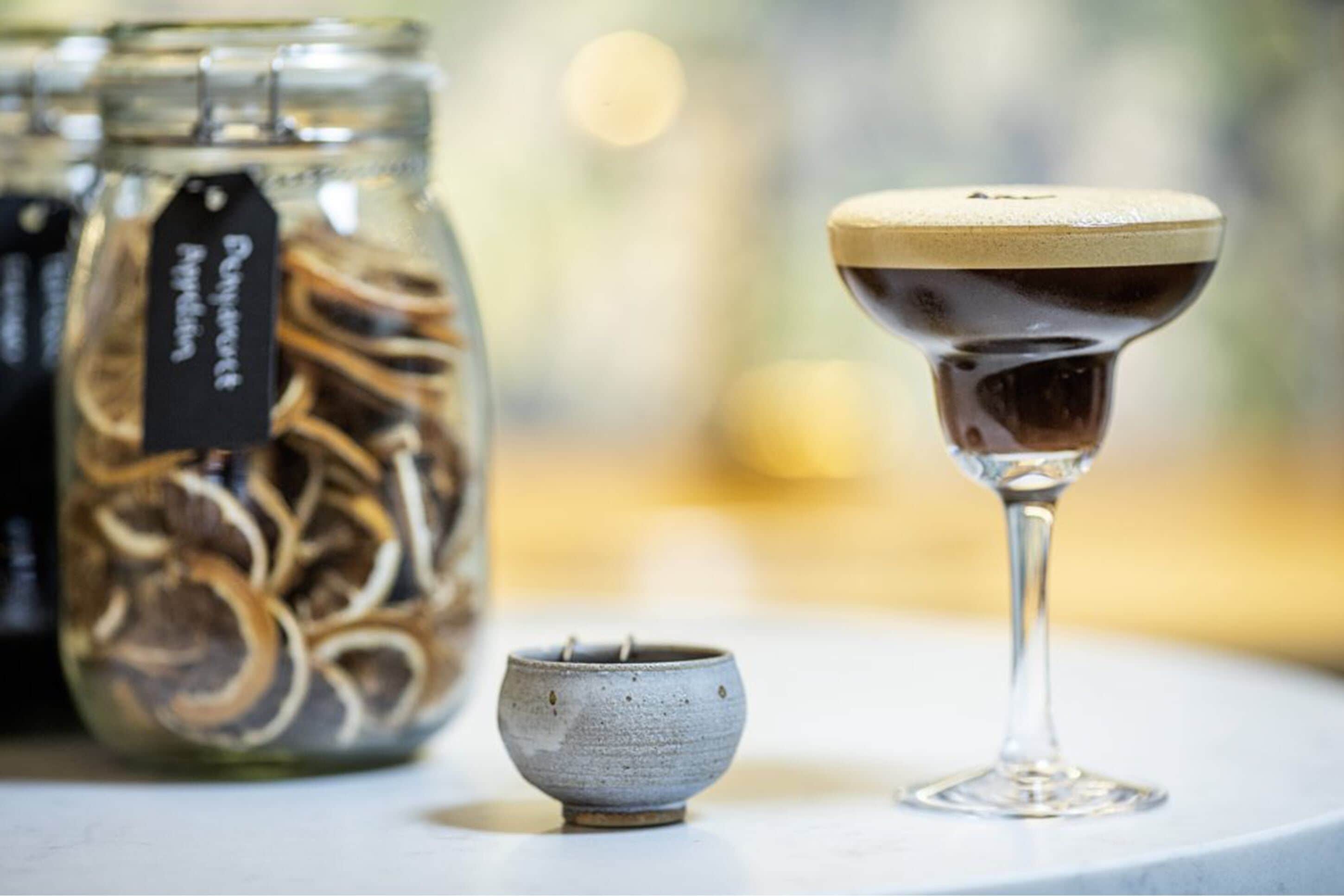 A glass of dark liquid with a frothy top, a small ceramic cup, and a jar with a tag labeled "Peppermint."