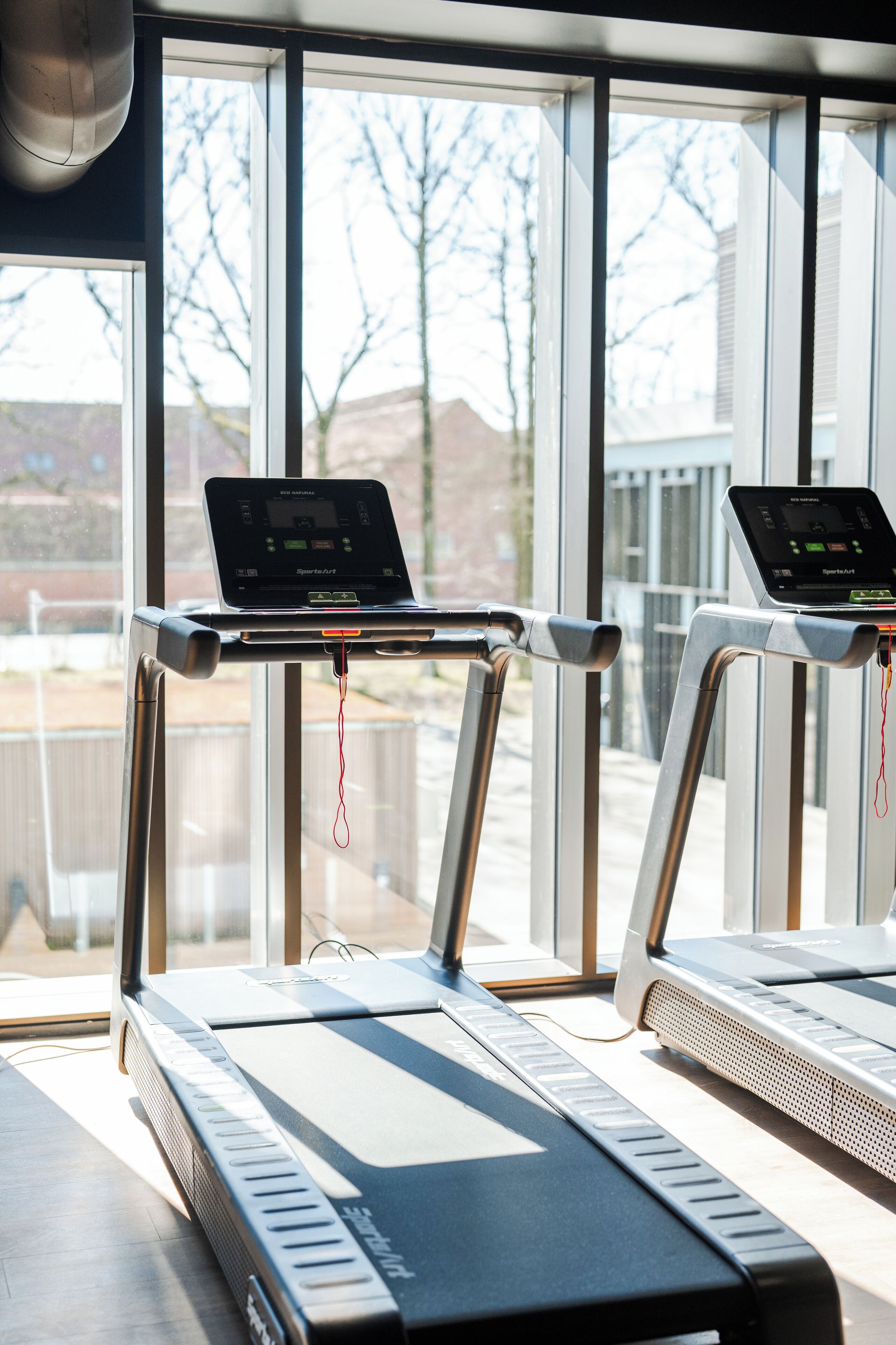 Ein Laufband mit digitaler Anzeige und einem Laptop darauf, vor großen Fenstern mit Blick auf Bäume und Gebäude platziert.