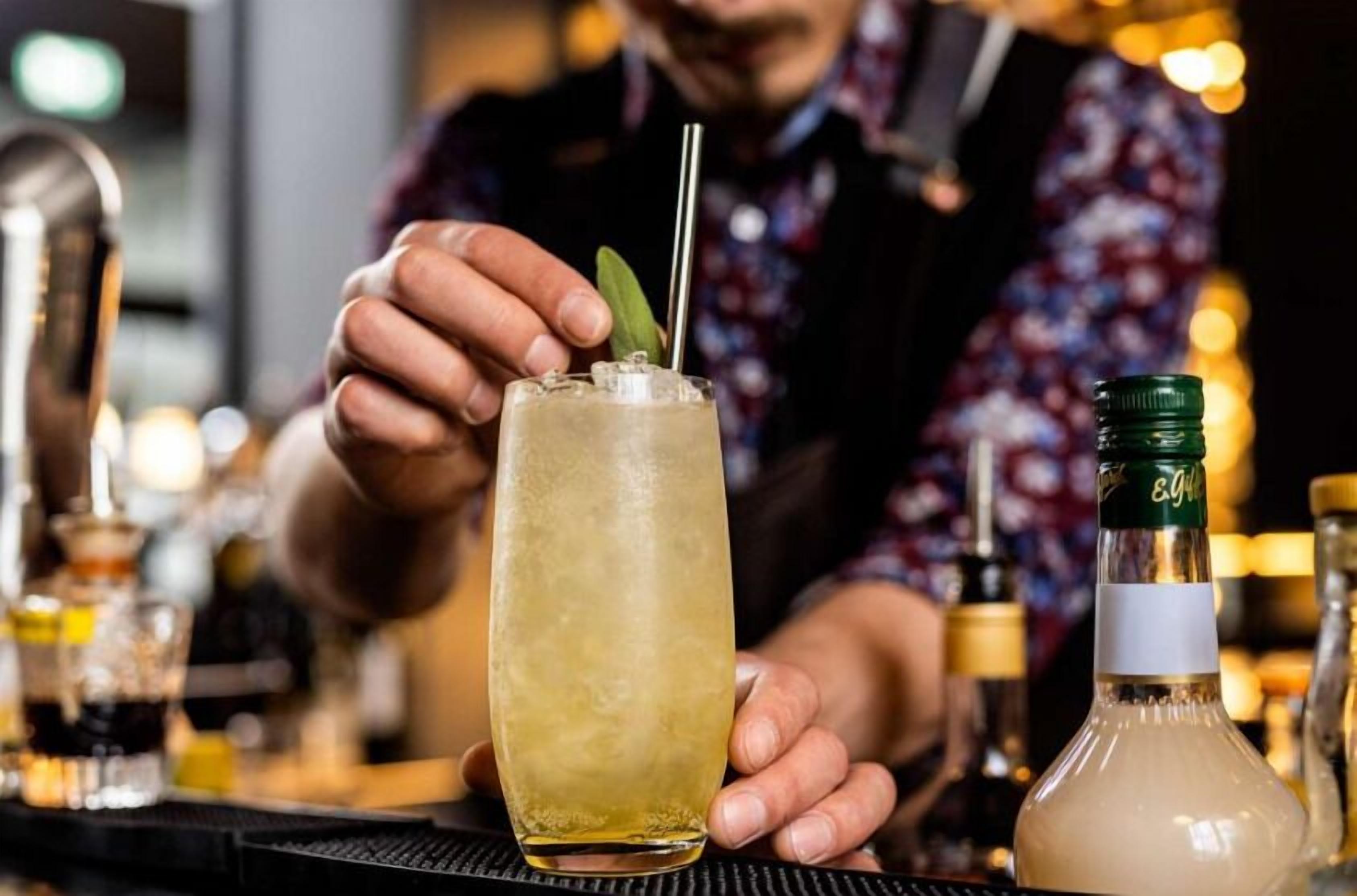 Un barman prépare un cocktail à l’aide d’un grand verre, d’une bouteille verte et d’une paille en métal.