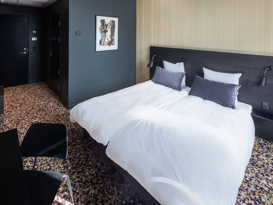 A hotel room with a large bed, a headboard, a wall-mounted lamp, and a carpeted floor.