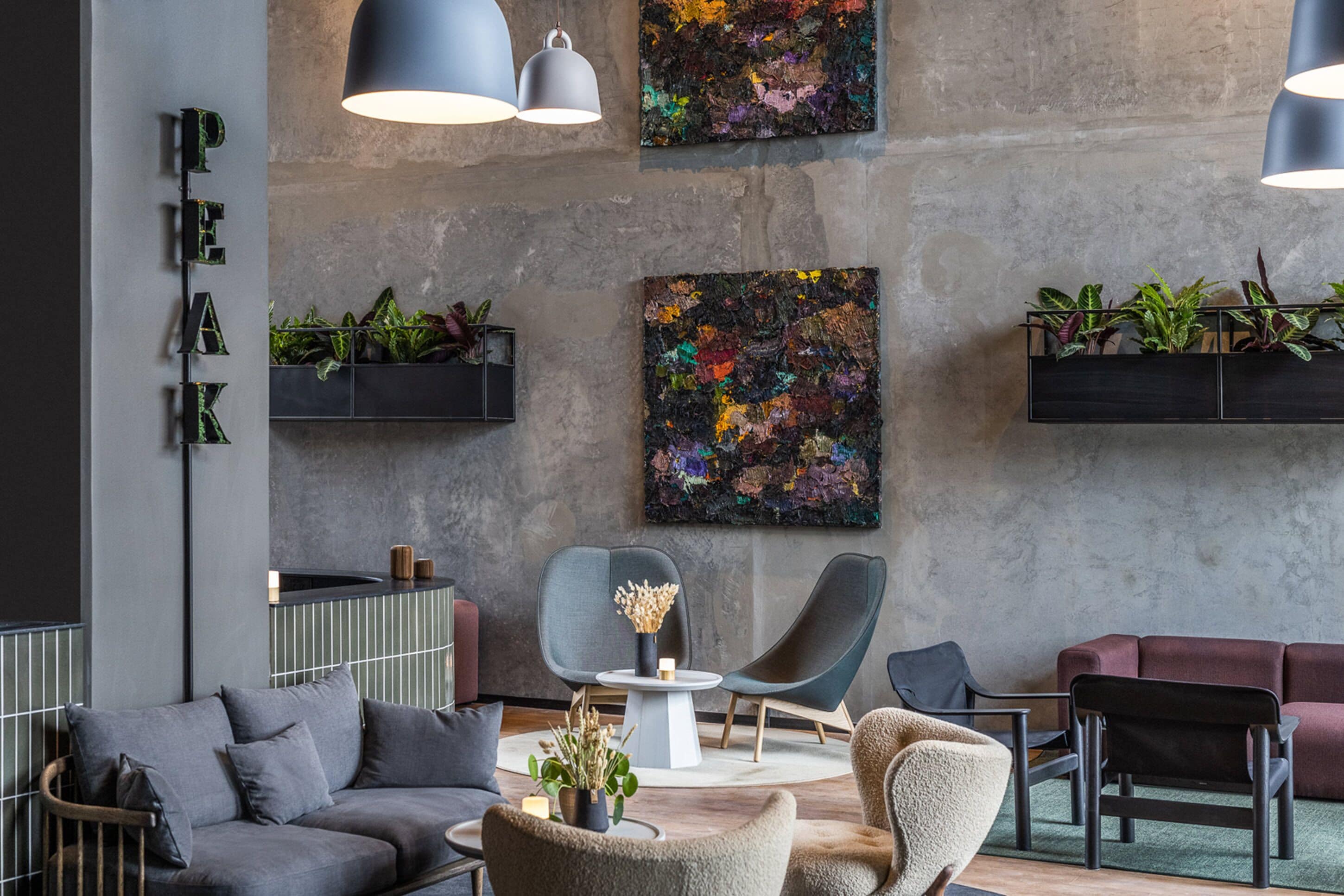 A modern lounge area with a grey sofa, armchairs, and a coffee table. There are two pendant lights, a black shelf with plants, and a colorful abstract painting on the wall.