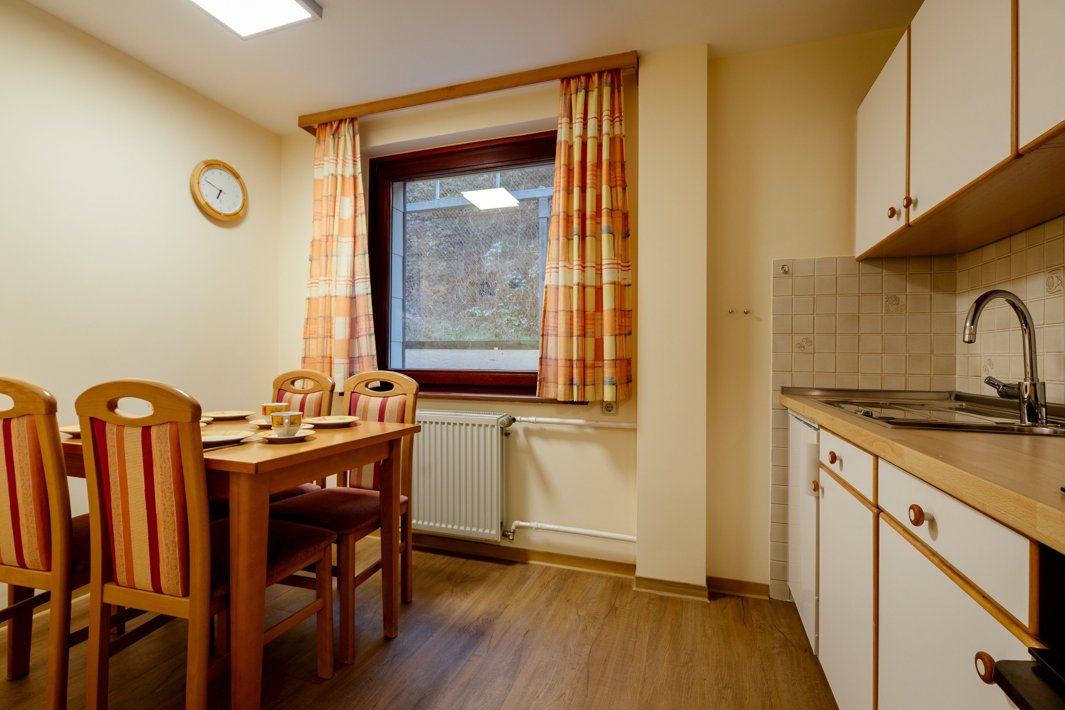A kitchen with wooden cabinets, a window with curtains, a radiator, and a dining table with chairs.