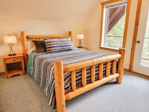 A wooden bed with a patterned bedspread, two bedside tables with lamps, and a window with a view of greenery.