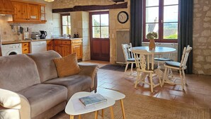 A cozy living room with a sofa, a coffee table, and a dining area with chairs and a table. There is a kitchen with wooden cabinets and a window with a view. - 2 Gites 8 Personnes Proche Beauval et Chenonceaux (Montrichard Val de Cher)