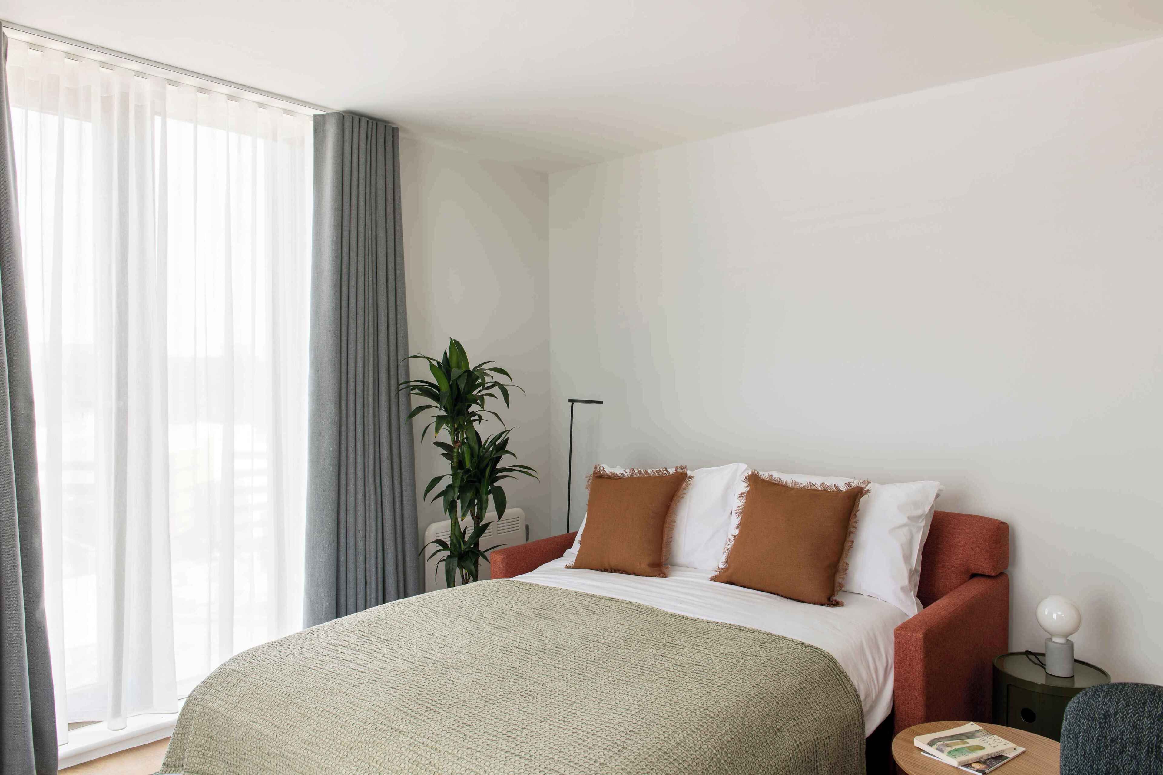 Un dormitorio con cama, mesita de noche, lámpara, planta y ventana con cortinas.