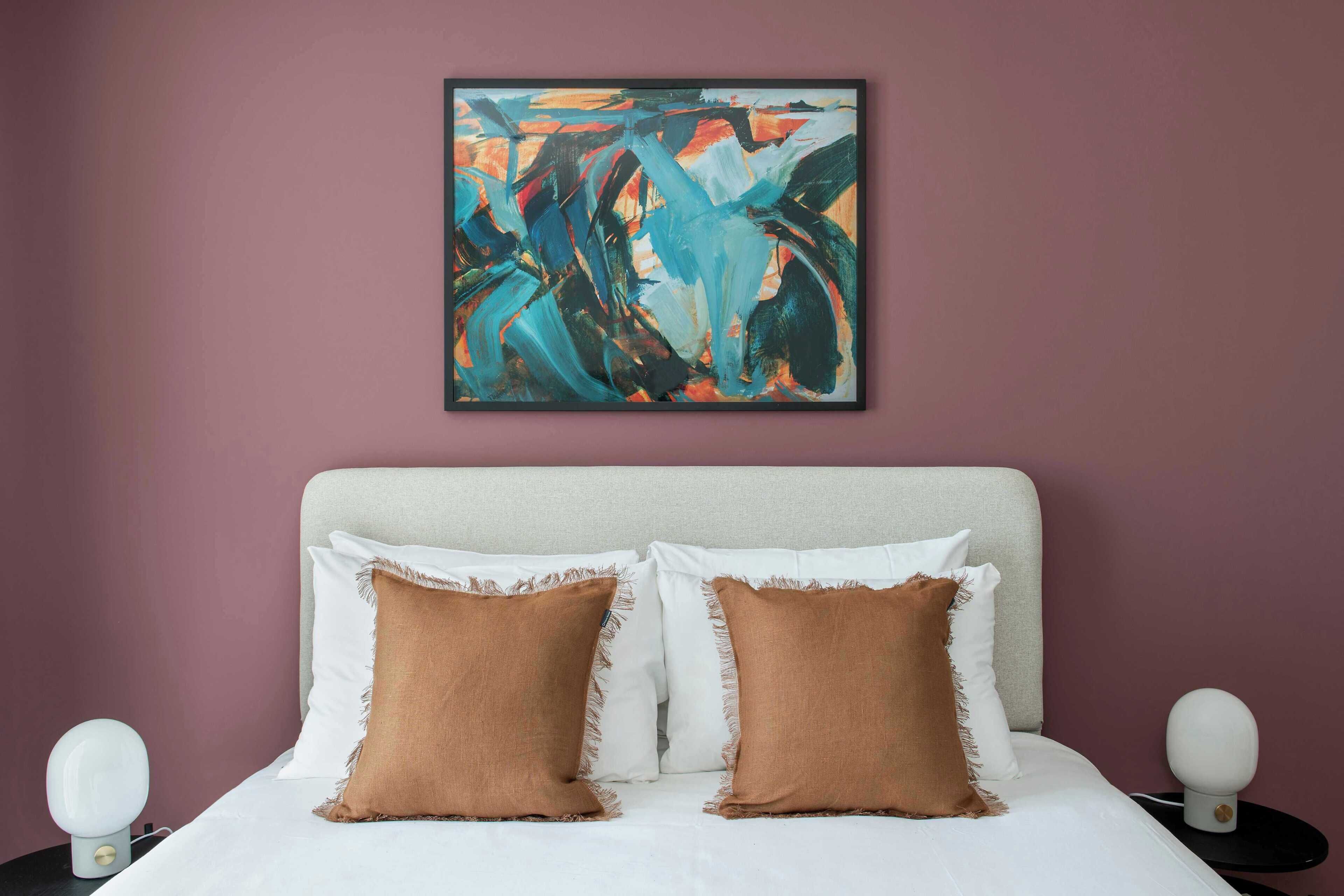 A bedroom with a bed, two pillows, a nightstand, a wall-mounted abstract painting, and a bedside lamp.