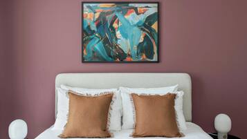 A bedroom with a bed, two pillows, a nightstand, a wall-mounted abstract painting, and a bedside lamp.