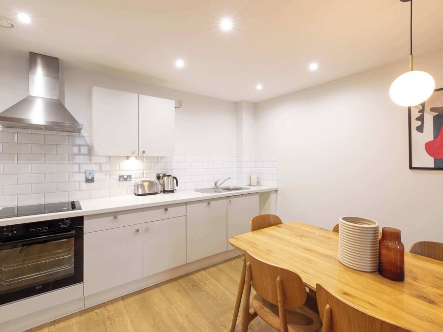 A modern kitchen with white cabinetry, a stainless steel range hood, a built-in oven, a sink, and a dining area with a wooden table and chairs.