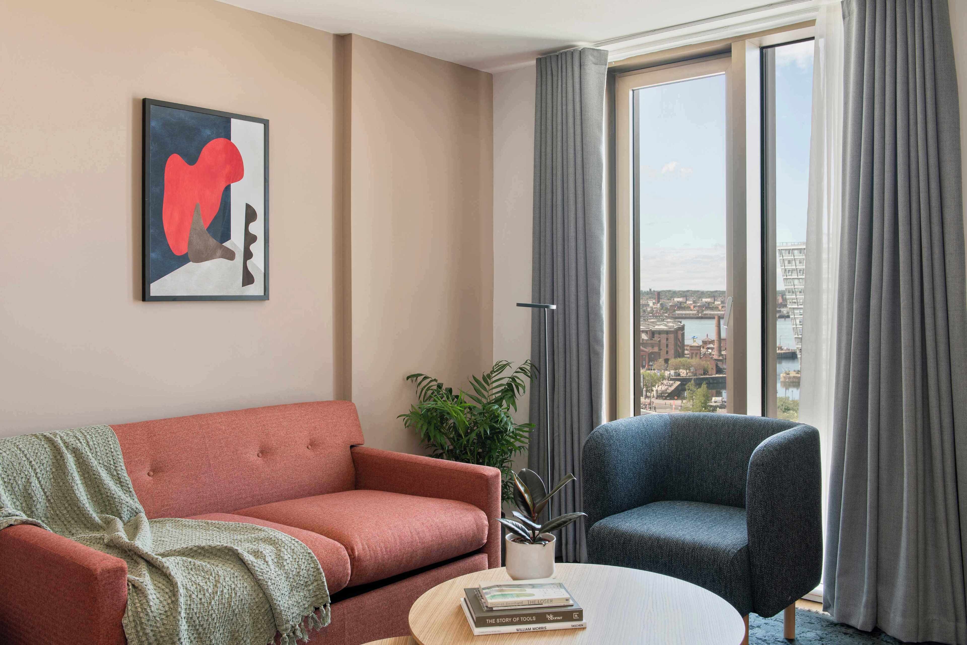 A modern living room with a red sofa, a blue armchair, a round coffee table, and a city view through large windows.