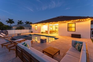 Un patio trasero con piscina, mobiliario exterior e iluminación colgante en la casa.