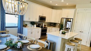 Una cocina moderna con armarios blancos, electrodomésticos de acero inoxidable y un comedor con mesas y sillas de madera.