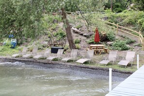 Ein Uferbereich mit Holzterrasse, Liegestühlen, Picknicktisch und einem roten Sonnenschirm.