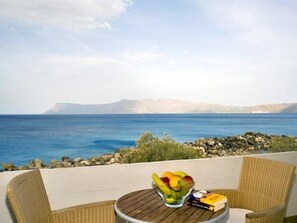 Un balcón con mobiliario de mimbre, un cuenco de vidrio con fruta y un libro, con vistas a la costa, un litoral rocoso y montañas distantes.