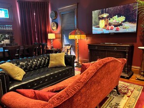 A living room with a black leather sofa, a red armchair, a television displaying a fruit scene, and a dining area in the background.