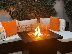 A fire pit table with a white cushioned sofa and orange cushions, surrounded by greenery.