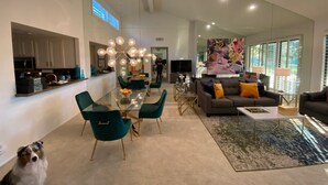 Een moderne woonkamer met een glazen eettafel, smaragdgroene stoelen, een grijze bank met oranje kussens, en een groot abstract schilderij aan de muur.