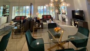 Een ruime woonkamer met een hoge plafond, een grote kroonluchter en een open haard. Er is een televisie aan de muur bevestigd en een eethoek met een glazen tafel en stoelen.