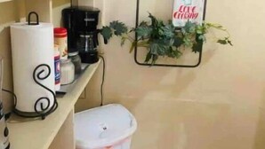 Een keuken met een koffiezetapparaat, een prullenbak en een plank met keukengerei.