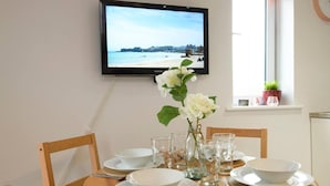 A dining area with a table set for four, a television mounted on the wall displaying a beach scene, and a window with a plant. - 1 bed Apartment Close to Football Grounds (Liverpool)