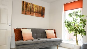 A grey sofa with two cushions, a painting of a forest, a window with a plant, and a door. - 1 bed Apartment Close to Football Grounds (Liverpool)