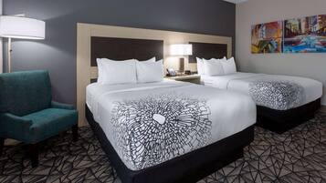 A hotel room with two beds, a teal armchair, a nightstand with a lamp, and a cityscape painting on the wall.