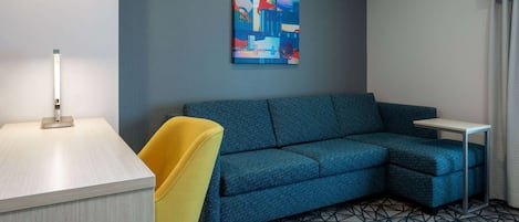 A modern hotel room with a blue sofa, a yellow armchair, a small table, and a geometric patterned floor.