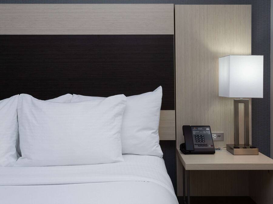 A hotel room with a bed, white pillows, a bedside table with a lamp, and a phone.