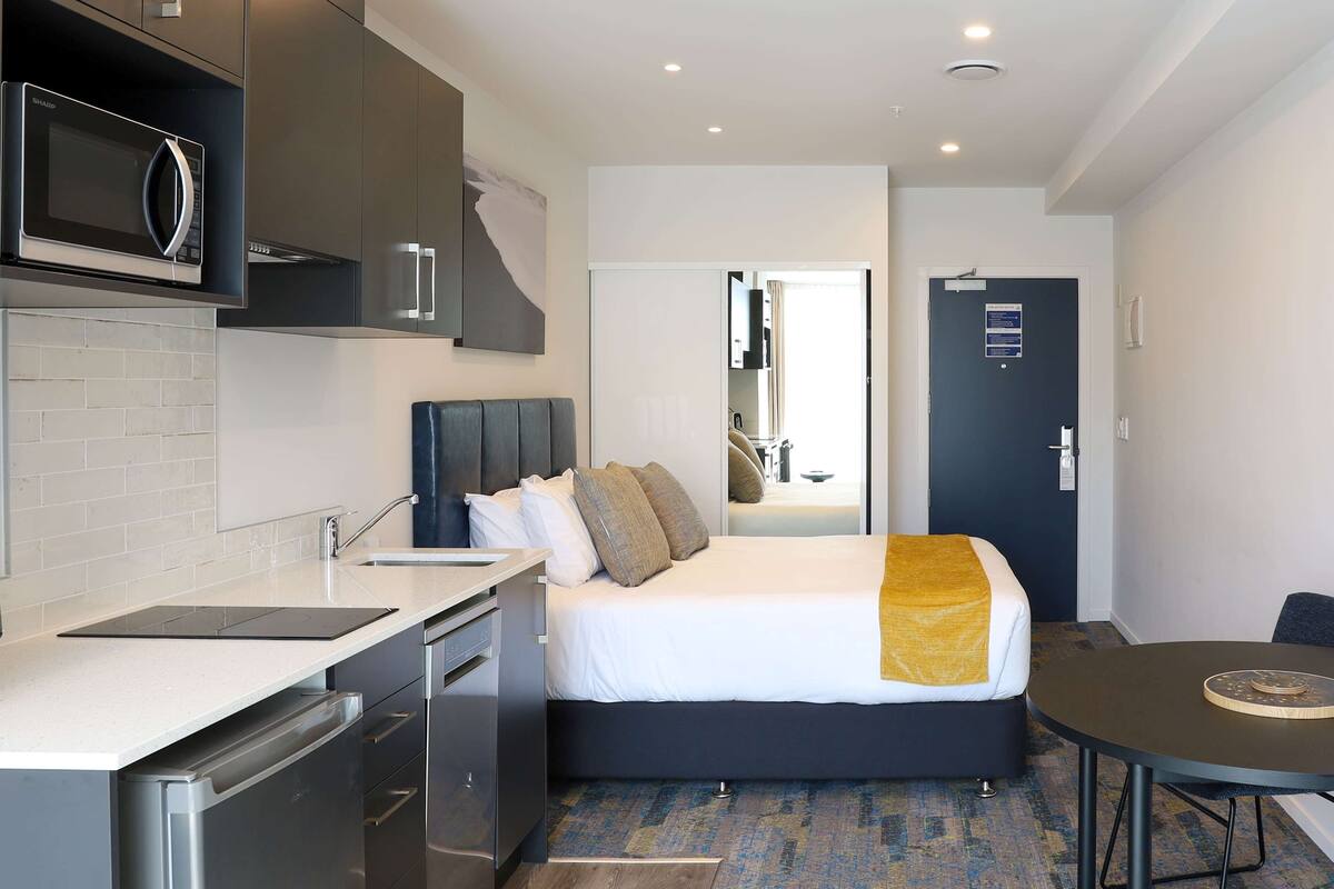 Ein modernes Hotelzimmer mit Küchenzeile, einem Bett mit Kopfteil, einem Nachttisch und einem runden Tisch.