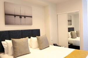 Camera d'albergo con un letto, cuscini, una testiera, un quadro raffigurante barche sull'acqua e uno specchio che riflette la stanza.