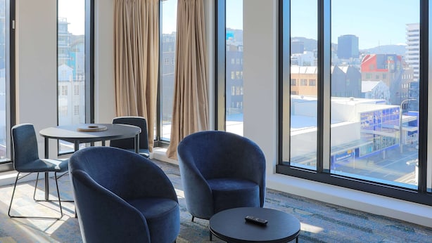 Une chambre d’hôtel moderne avec de grandes fenêtres, une table ronde, deux fauteuils bleus et une télécommande posée sur la table.