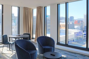 Een moderne hotelkamer met grote ramen, een ronde tafel, twee blauwe fauteuils en een afstandsbediening op de tafel.