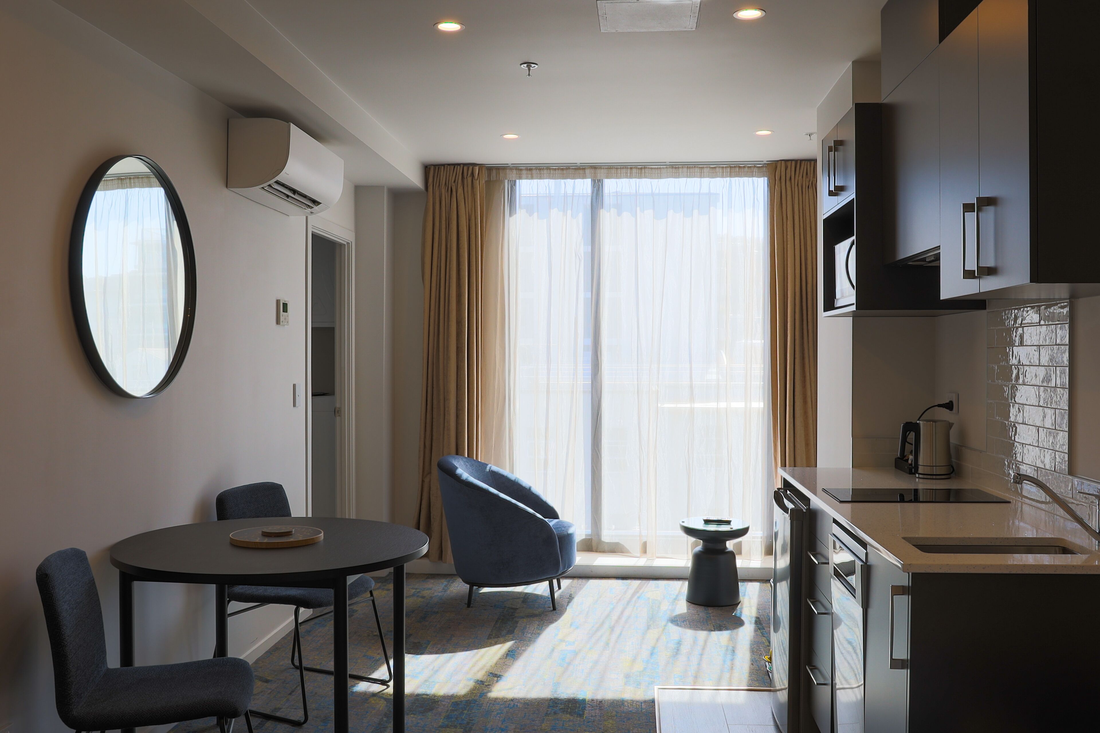 Una habitación de hotel moderna con zona de comedor, sala de estar, kitchenette y un ventanal con cortinas.