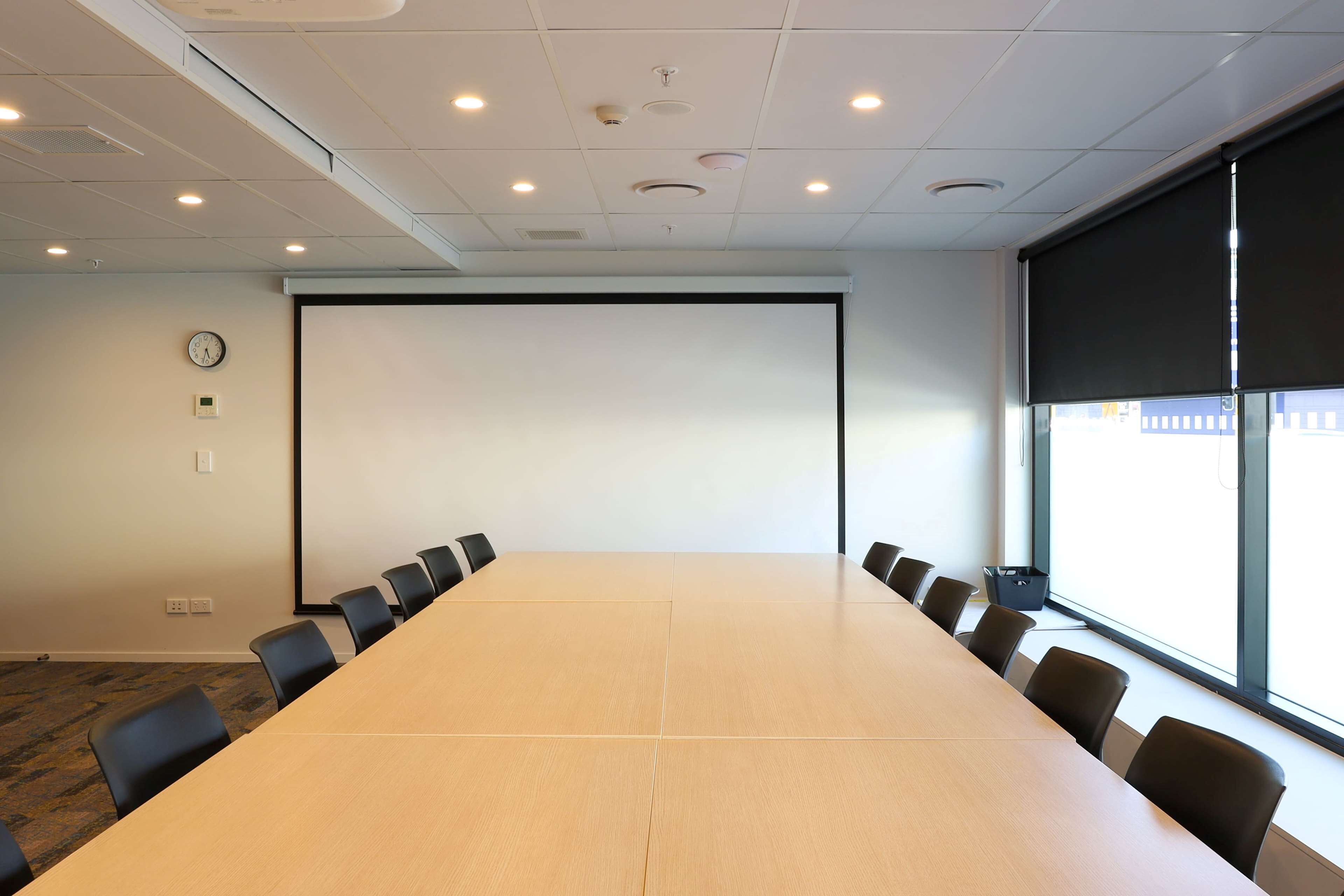 A conference room with a long table, chairs, a projector screen, a clock, and a window with blinds.