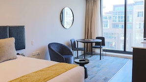 Une chambre d’hôtel avec un lit, un miroir rond, une petite table avec des chaises et une vue sur les bâtiments à travers de grandes fenêtres.