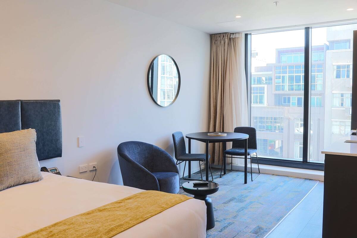 Ein Hotelzimmer mit einem Bett, einem runden Spiegel, einem kleinen Tisch mit Stühlen und Blick auf Gebäude durch große Fenster.