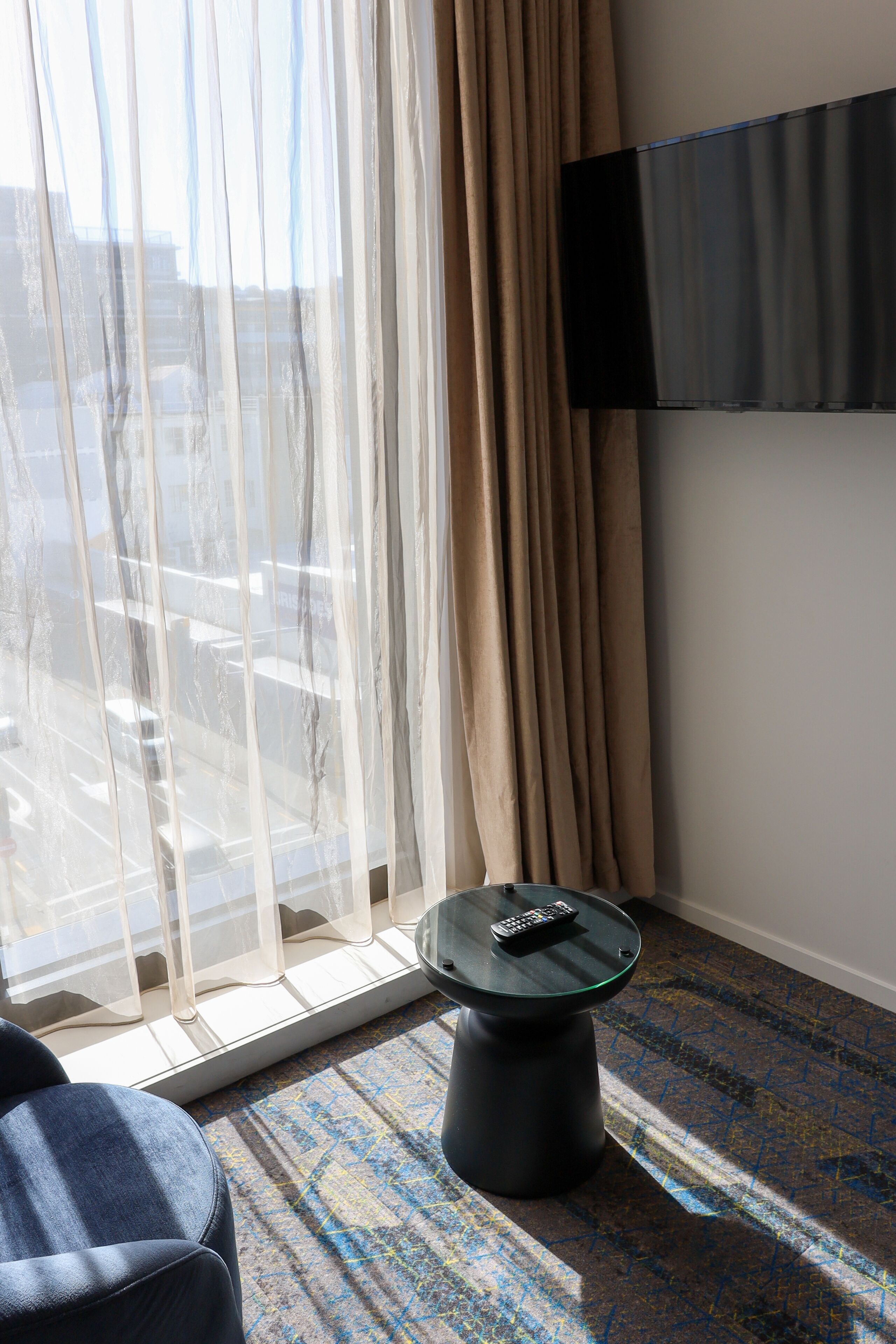 A hotel room with a large window, a remote control on a small table, and a flat-screen TV mounted on the wall.