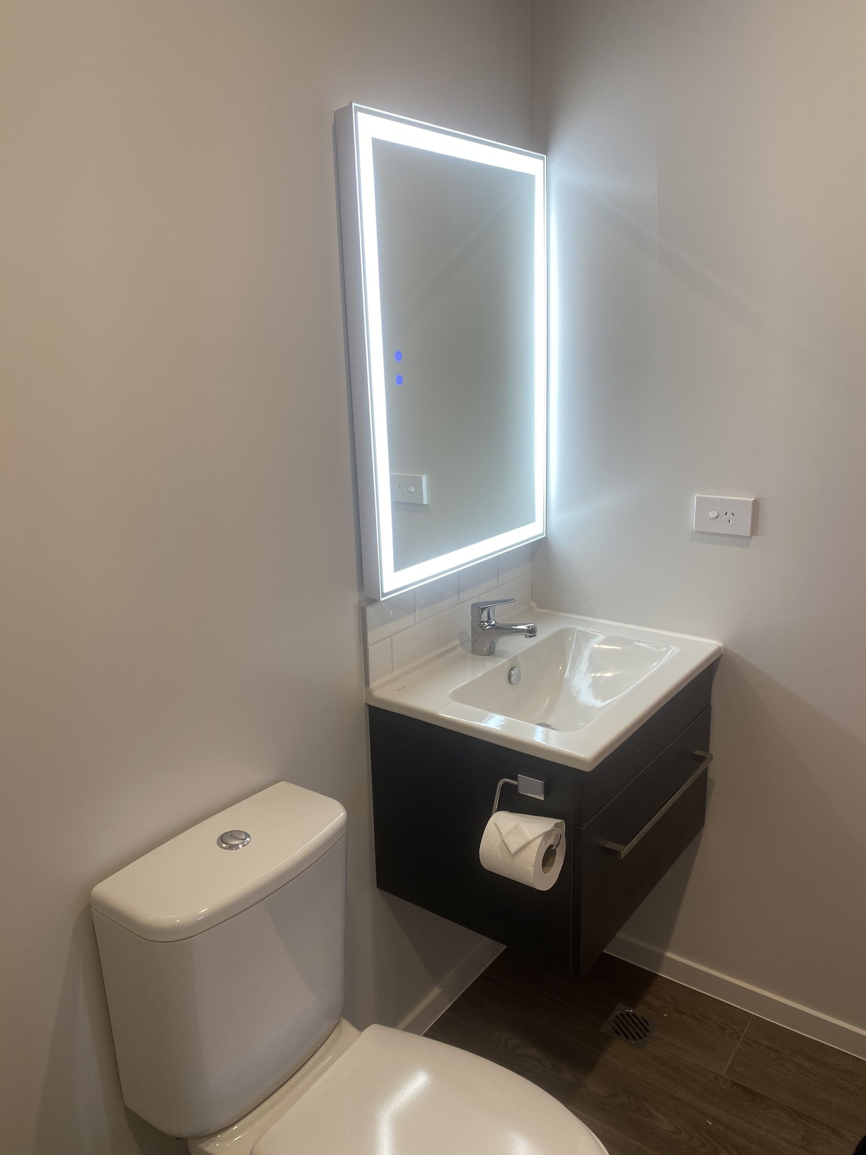 Un baño moderno con inodoro blanco, lavamanos con espejo y un mueble bajo.
