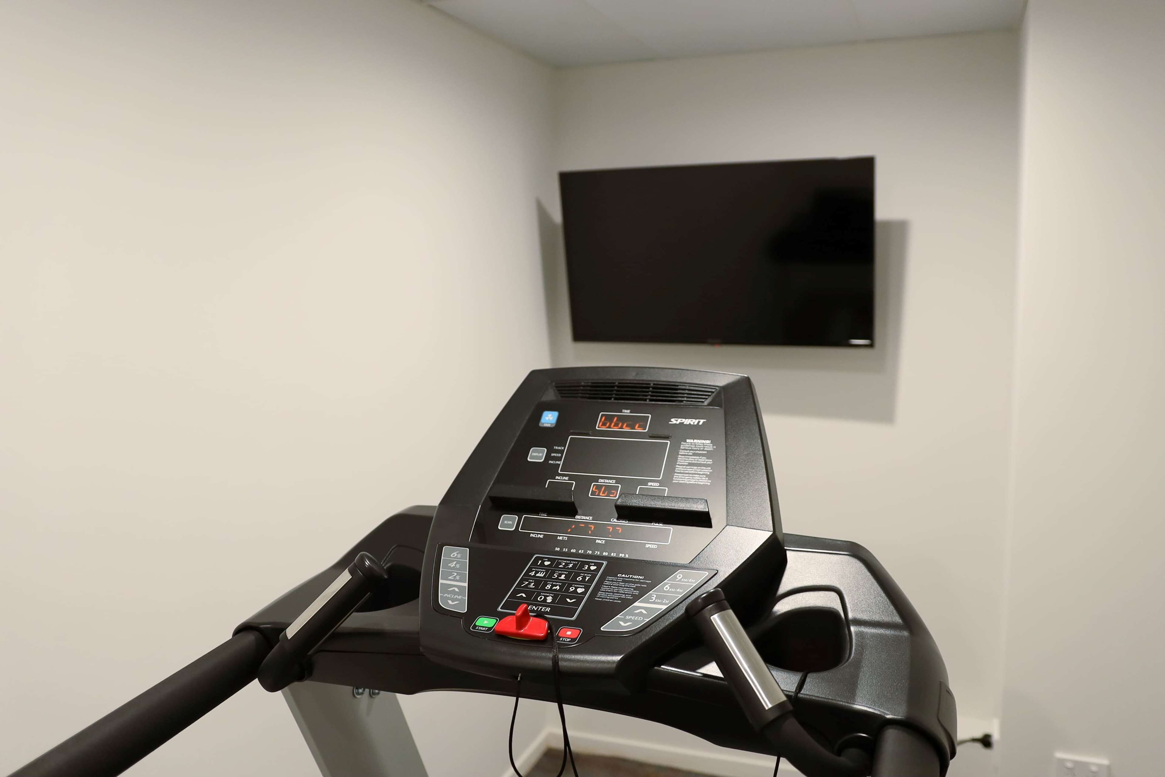 A treadmill with a digital display and control panel, a wall-mounted TV, and a plain white wall.