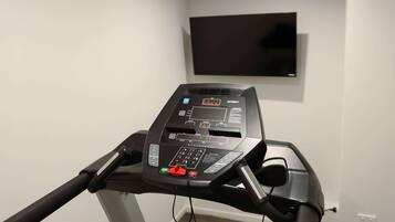 A treadmill with a digital display and control panel, a wall-mounted TV, and a plain white wall.