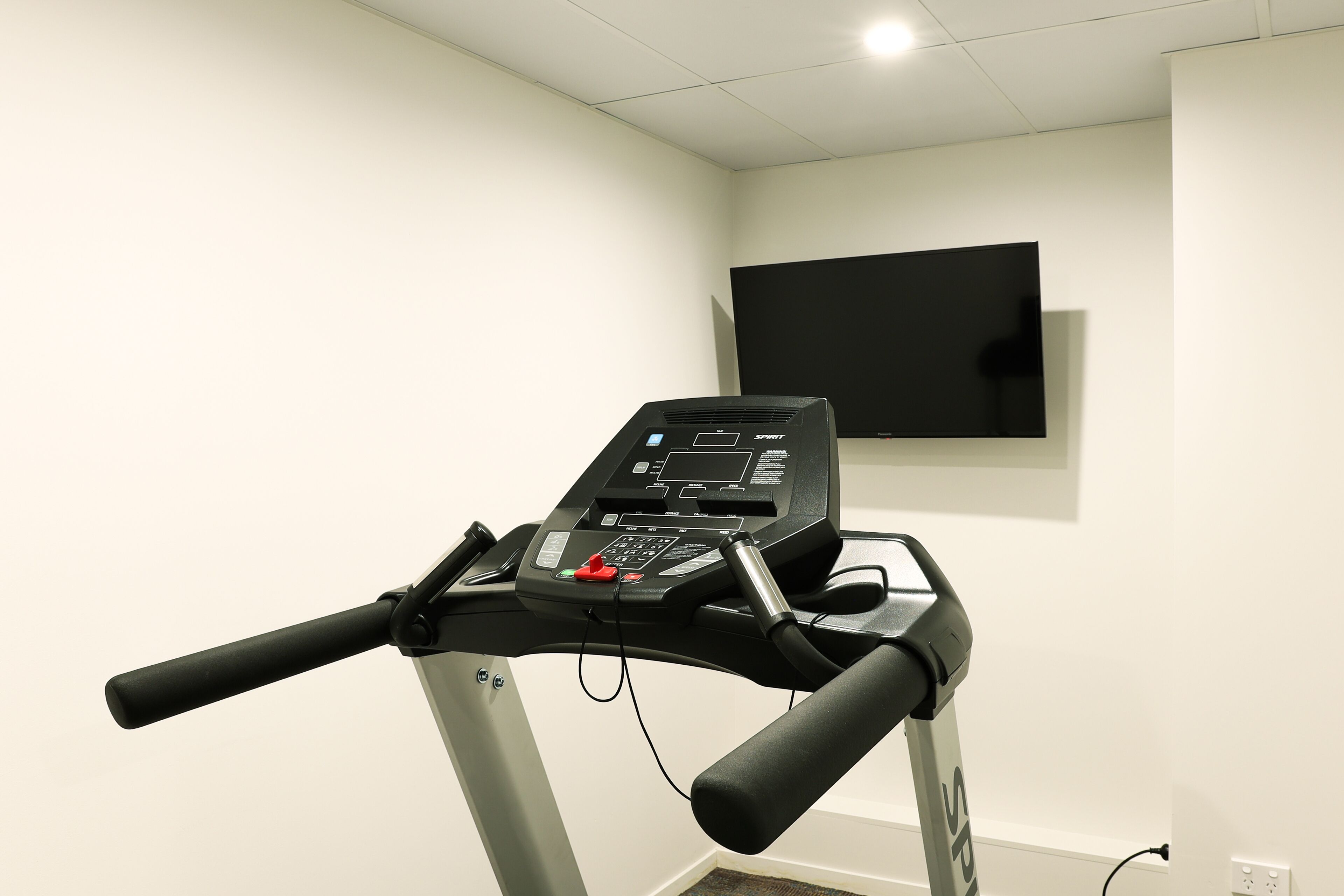 A treadmill with a digital display and control panel, a wall-mounted TV, and a power outlet on the wall.