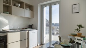A modern kitchen with a view of a cityscape through large windows, a dining area with a table set for two, and a small balcony with a potted plant.