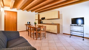 A living room with a sofa, wooden dining table, chairs, a TV on a stand, and a kitchen area with cabinets and a sink. - Agriturismo Colombare Teboi (Colombare)