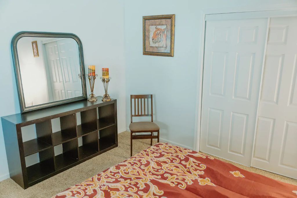 Ein Zimmer mit einem großen Spiegel, einem Holzstuhl, einem gemusterten Teppich, einem Beistelltisch mit Kerzenhalter und einer Tür.